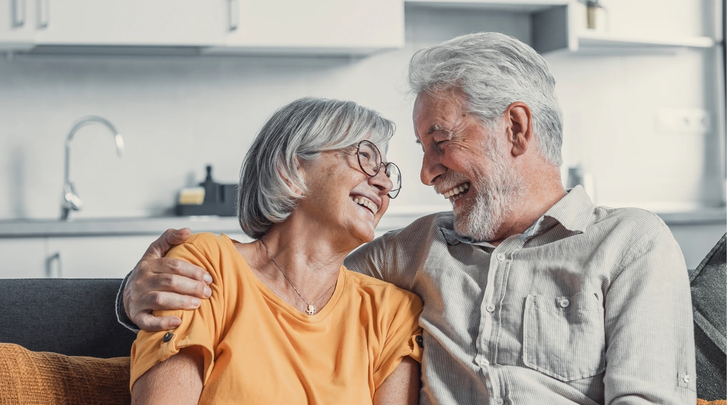 Two seniors smiling while sitting on a couch and looking at each other